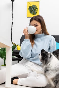 A teenager sitting on the floor relaxes drinking a cup of fresh coffee and her faithful companion dog is lying next to her. Purebred Border Collie dog in shades of white and black, and long and fine hair. Living room in a modern styling.
