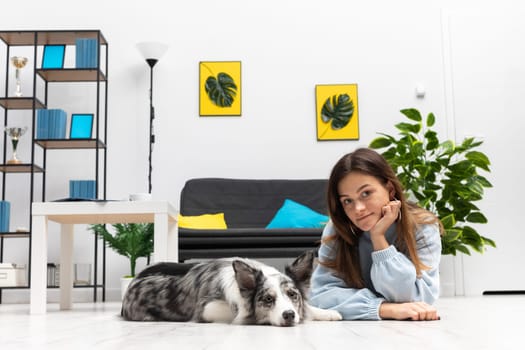 The dog sitter lies with him on the floor of the owl house in the living room and rests during a break from training. Purebred Border Collie dog in shades of white and black, and long and fine hair. Living room in a modern styling.