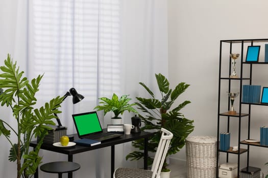 In the privacy of your home, due to the need to work remotely, a table with a laptop, calendar and lamp was placed. A bright living room in a modern style with a preferable space. Green screen of the monitor.
