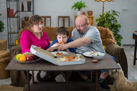 A shaggy dog watches when he too gets his slice. Mother, Father and son help themselves to hot and fragrant slices of pizza from a box.