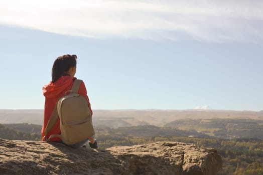 A girl tourist with a backpack sits on the top of a mountain and enjoys the beautiful views. A walking woman in an orange jacket rests on a rock, looking at the beautiful sunny landscape of the Caucasus Mountains