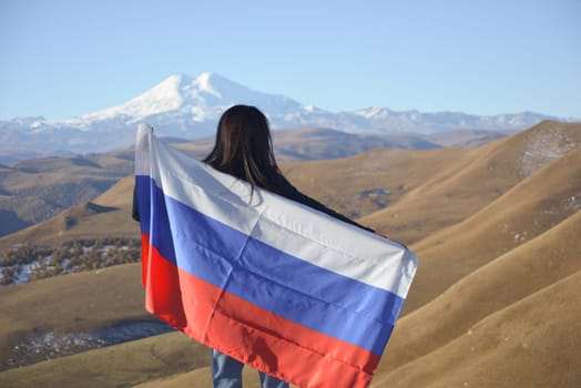 A young brunette woman stands against the backdrop of the snow-capped Mount Elbrus, looking at the beauty of the mountains, holding a Russian flag in her hands. Tricolor against the backdrop of snow-capped Mount Elbrus.