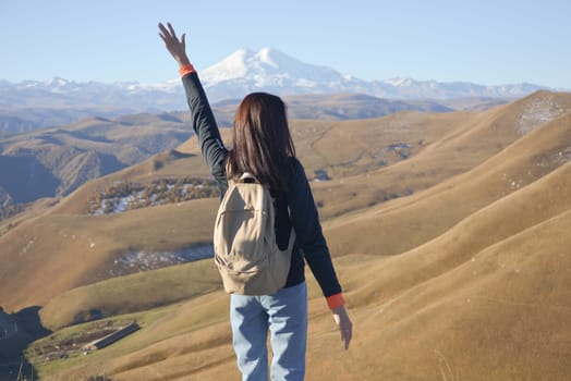 A young brunette woman with a backpack stands against the background of snow-capped Mount Elbrus and looks towards the mountains