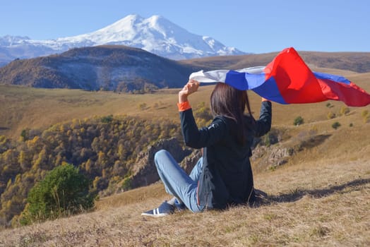A young brunette woman sits on a stone and looks at the Caucasus Mountains around, a Russian flag flutters in her hands. Tricolor against the backdrop of snow-capped Mount Elbrus.