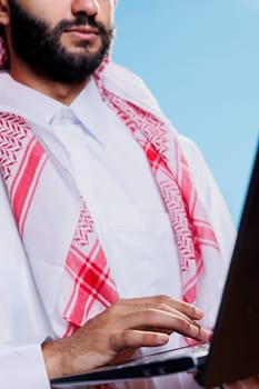 Arab man in thobe and headscarf holding laptop and writing message on keyboard closeup on hand. Muslim person wearing traditional clothes using portable computer, texting and typing