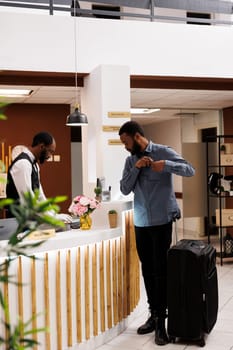 Young African American man tourist arriving to hotel, standing alone with suitcase at reception counter. Black millennial guy traveler checking in at front desk, waiting for room key
