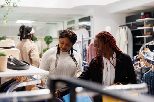 African american women friends browsing apparel and accessories in clothing store. Girlfriends checking garment for sale hanging on rack and shopping together in mall boutique