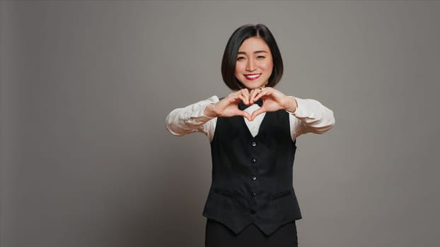 Hotel administrator shows heart shape symbol on camera, expressing romantic gesture and being flirty. Asian receptionist presenting true honest feelings, does romance sign in studio. Camera A.