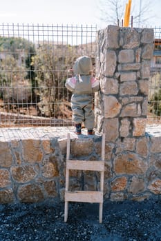 Small child in a hooded overall stands near a metal fence and looks out into the park. Back view. High quality photo