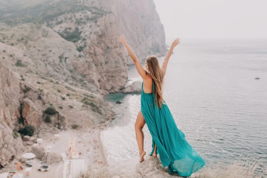 Side view a Young beautiful sensual woman in a mint long dress posing on a volcanic rock high above the sea during sunset. Girl on the nature on overcast sky background. Fashion photo