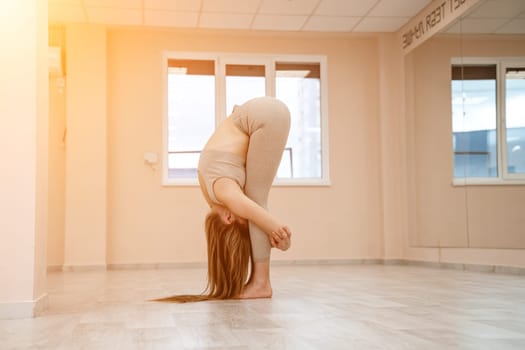 Young woman doing yoga in the gym. A girl with a long braid and in a beige tracksuit stands in a stork pose on a pink carpet. A woman performs padahstasana