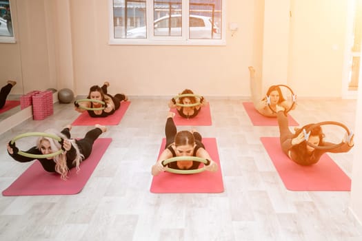 A group of six athletic women doing pilates or yoga on pink mats in front of a window in a beige loft studio interior. Teamwork, good mood and healthy lifestyle concept