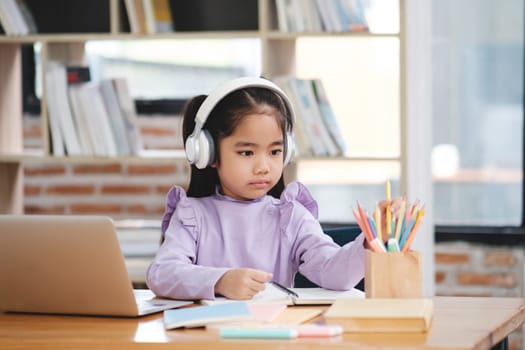 A young girl wearing headphones is sitting at a desk with a laptop and a box of colored pencils. She is focused on her work, possibly studying or doing homework. The scene suggests a quiet