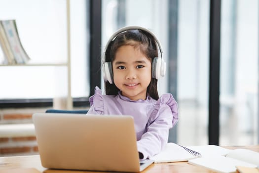 A young girl is sitting at a desk with a laptop and a notebook. She is wearing headphones and smiling. The scene suggests that she is engaged in some form of learning or studying