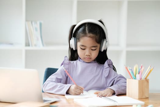 A young girl is sitting at a desk with a laptop and a notebook. She is wearing headphones and writing in her notebook
