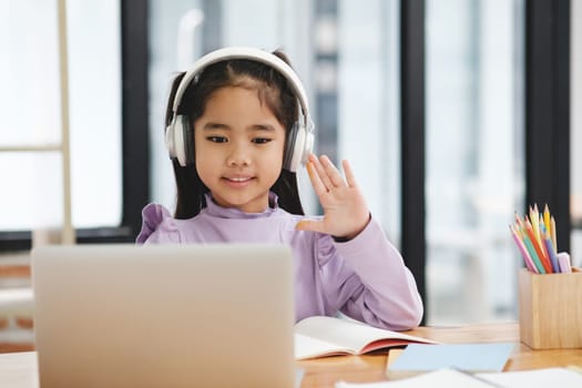 A young girl wearing headphones is waving at the camera while sitting at a desk with a laptop and a book