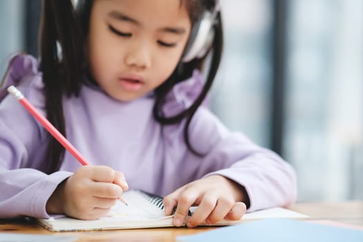A young girl is writing in a notebook with a pencil. She is wearing headphones and she is focused on her work. The scene suggests that she is engaged in a task that requires concentration