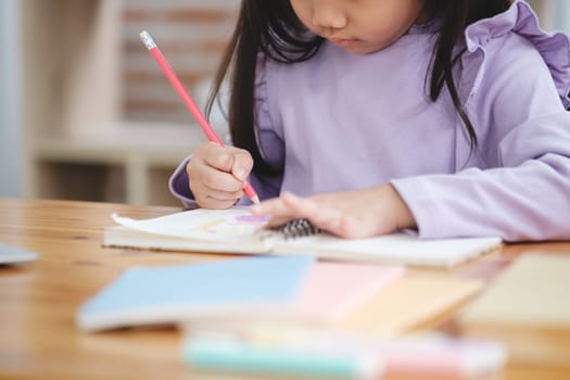 A young girl is writing with a red pencil on a piece of paper. She is sitting at a desk with a laptop and a stack of books nearby