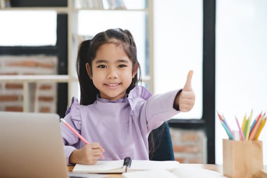 A young girl is sitting at a desk with a laptop and a notebook. She is smiling and giving a thumbs up. The scene suggests that she is happy and enjoying her work or activity