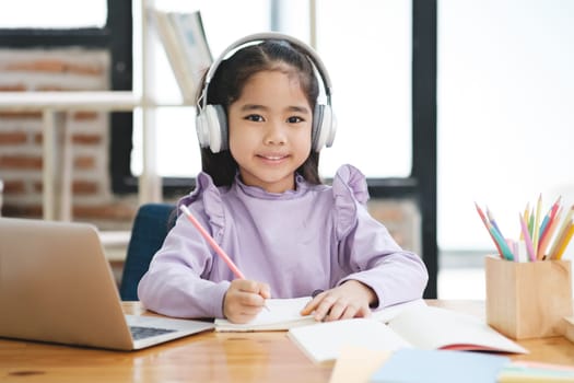 A young girl is sitting at a desk with a laptop and a notebook. She is wearing headphones and smiling. The scene suggests that she is engaged in some form of learning or studying