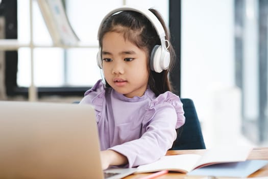 A young girl is sitting at a desk with a laptop and a notebook. She is wearing headphones and she is focused on her work. The scene suggests that she is engaged in a task that requires concentration