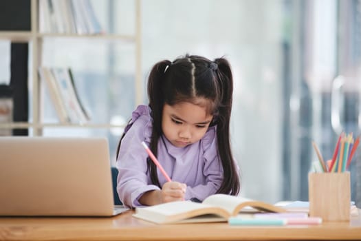 A young girl is sitting at a desk with a laptop and a book. She is writing with a pencil and she is focused on her work. The scene suggests a quiet and studious atmosphere