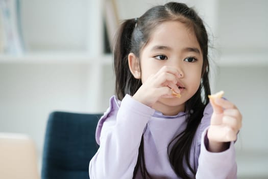 A young girl is eating a snack while sitting in a chair. She is wearing a purple shirt and has her hair in pigtails