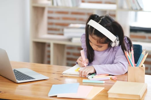 A young girl is sitting at a desk with a laptop and a notebook. She is drawing with markers and wearing headphones. The scene suggests that she is working on a project or assignment