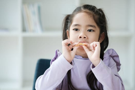 A young girl is eating a snack while sitting in a chair. She is wearing a purple shirt and has her hands in her mouth