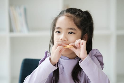 A young girl is eating a piece of food while sitting in a room with bookshelves. The scene is calm and peaceful, with the girl focused on her food