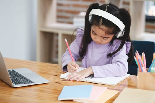 A young girl is sitting at a desk with a laptop and a notebook. She is writing in her notebook with a pencil. The scene suggests that she is working on a school assignment or studying for an exam