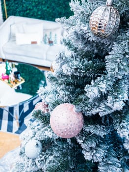 A snow-dusted Christmas tree adorned with glittering ornaments stands in the foreground. Behind it, a stylish living space features a white couch, a verdant green wall, and playful seasonal decor atop a sleek coffee table.