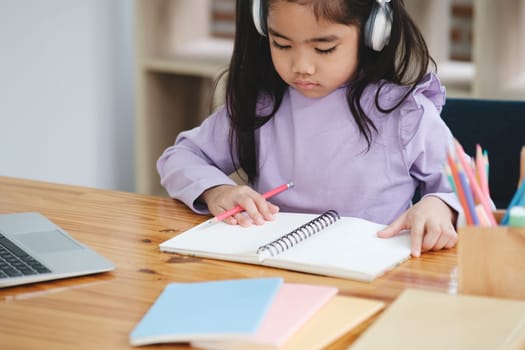A young girl is sitting at a desk with a notebook and a laptop. She is wearing headphones and she is focused on her work
