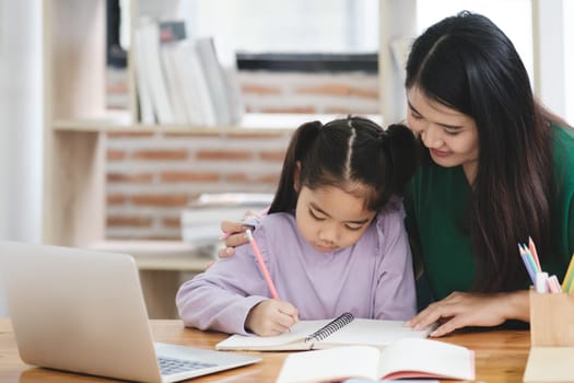 A woman is helping a young girl with her homework. The girl is wearing a purple shirt and is sitting at a desk with a laptop and a notebook