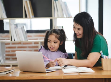 A woman is helping a young girl with her homework. The girl is using a laptop and a pencil. The woman is smiling and she is patient and supportive. The scene suggests a positive