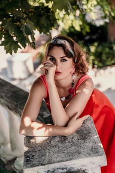 A pretty woman in a red silk dress and a bandage on her head smiles against the background of the leaves of a tree. She is leaning on the coop and looking into the camera. Vertical photo