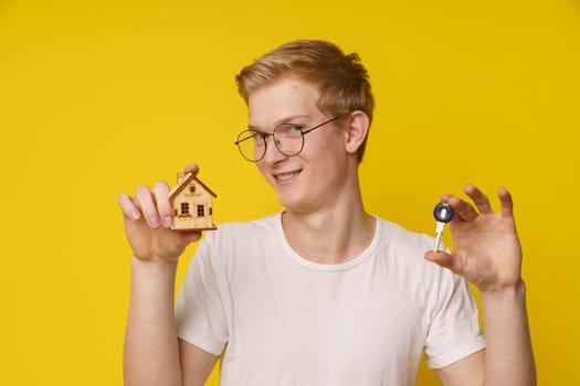 Young man with smile holds house model and home key in his hand, against yellow background with plenty of copy space. Concept of real estate, homeownership, rental, investment, or housing market. High quality photo