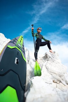 beautiful sup surfer girl in a wetsuit on the ocean shore against a beautiful blue sky poses sexually with a paddle