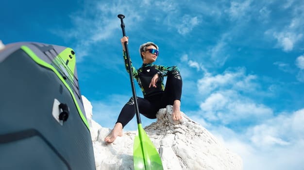 beautiful sup surfer girl in a wetsuit on the ocean shore against a beautiful blue sky poses sexually with a paddle