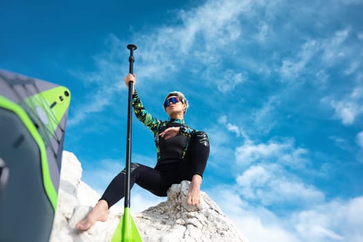 beautiful sup surfer girl in a wetsuit on the ocean shore against a beautiful blue sky poses sexually with a paddle