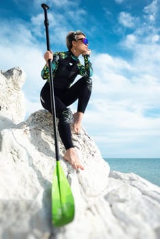 beautiful sup surfer girl in a wetsuit on the ocean shore against a beautiful blue sky poses sexually with a paddle