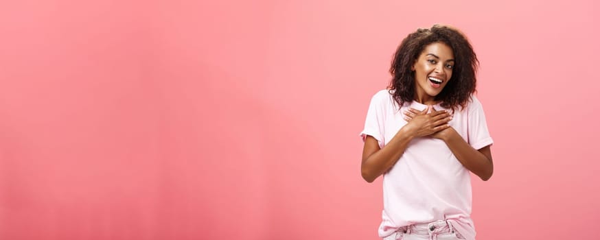 Portrait of charming delighted african american woman with curly haircut holding palms on heart pleased and grateful thanking friend for help smiling happily and thankful at camera over pink wall. Lifestyle.