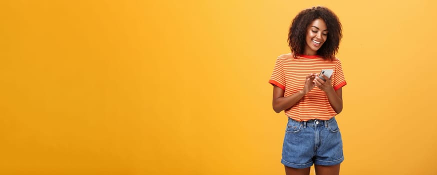 Stylish carefree girl texting friend come over standing pleased over orange wall in stylish denim shorts typing message or scrolling news in internet via smartphone gazing at device screen with smile. Lifestyle.