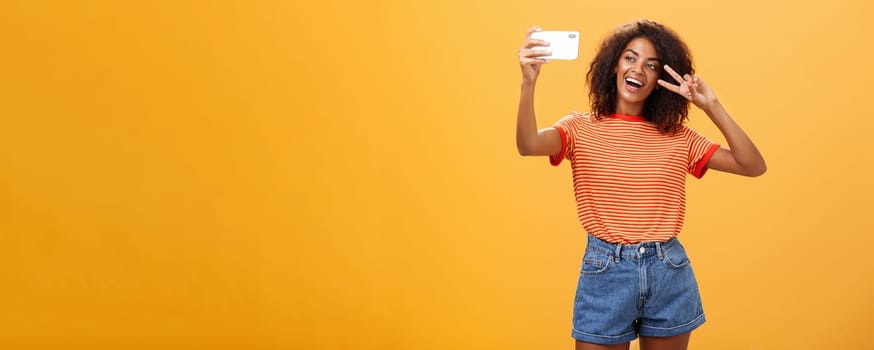 Girl making video vlog with brand new smartphone posting in internet trying become famous standing over orange background posing for selfie gazing at gadget screen showing peace or victory gesture. Copy space