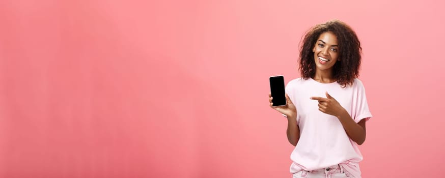Suggest this model of phone. Happy charming african american curly-haired woman in stylish outfit holding smarpthone and pointing at device screen while recommending cool app over pink wall.