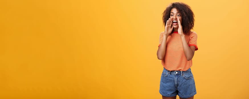 Girl calling coworker help out turning left with carefree joyful look holding hands near opened mouth to shout louder asking friend come closer while yelling on distance over orange background. Body language and lifestyle concept