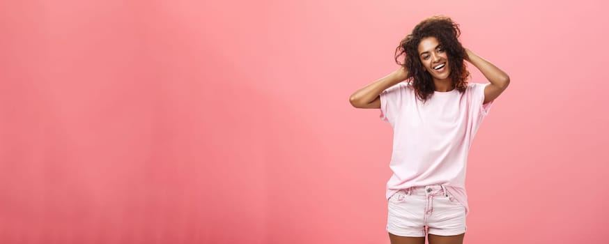 Girl enjoying nice weather chilling making photos for social network touching curly hair gently and carefree smiling broadly at camera standing over pink background happy and chill. Emotions and people concept