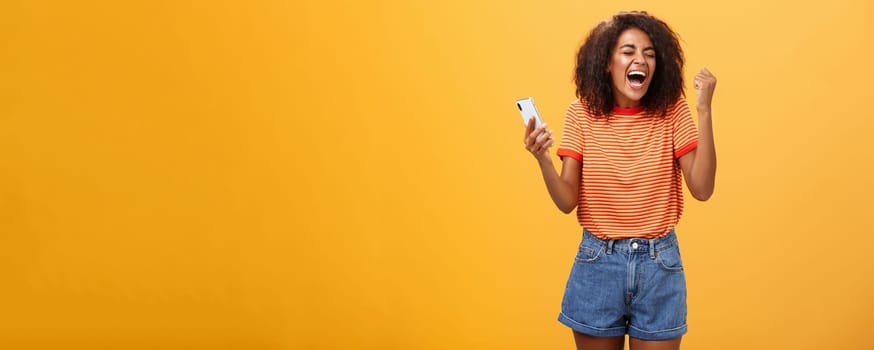 Portrait of ambitious happy young african american girl yelling from happiness and triumph clenching fist in joy and celebration feeling excited and relieved holding smartphone over orange wall. Lifestyle.
