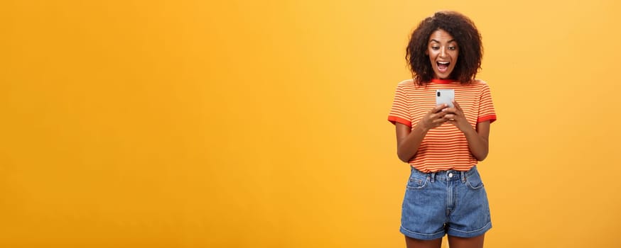 Girl expressing excitement and joy receiving awesome invitation via messages yelling from delight and happiness looking at smartphone screen impressed and thrilled over orange background.