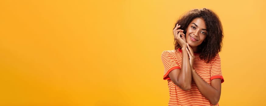 Waist-up shot of sensual feminine and flirty dark-skinned woman in trendy t-shirt holding arms near face smiling tenderly and gentle touching soft skin delighted and pleased over grey wall. Skincare, cosmetology and people concept